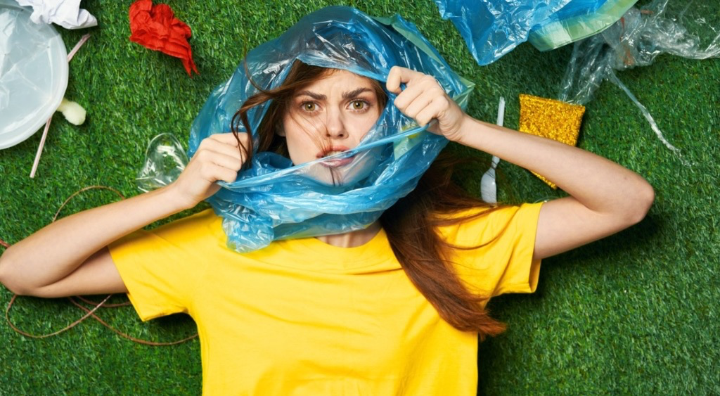 Une femme allongée sur l'herbe avec un sac plastique bleu autour de la tête, entourée de déchets plastiques